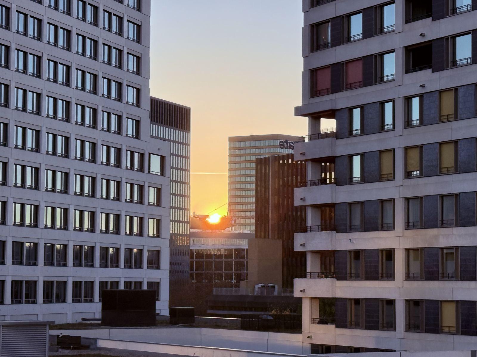 High-rise apartment and office buildings framing a low sun at dusk.