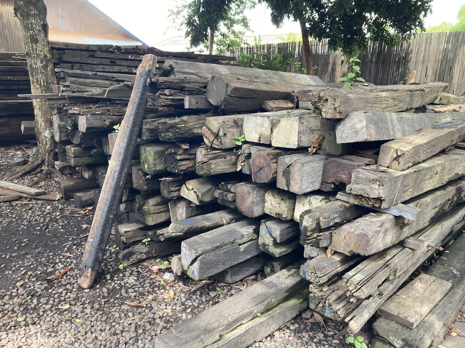 A yard stacked with salvaged ulin beams at Kaltimber in Bali, weathered grey and green with moss, chalk-marked with their dimensions and still carrying old bolt holes