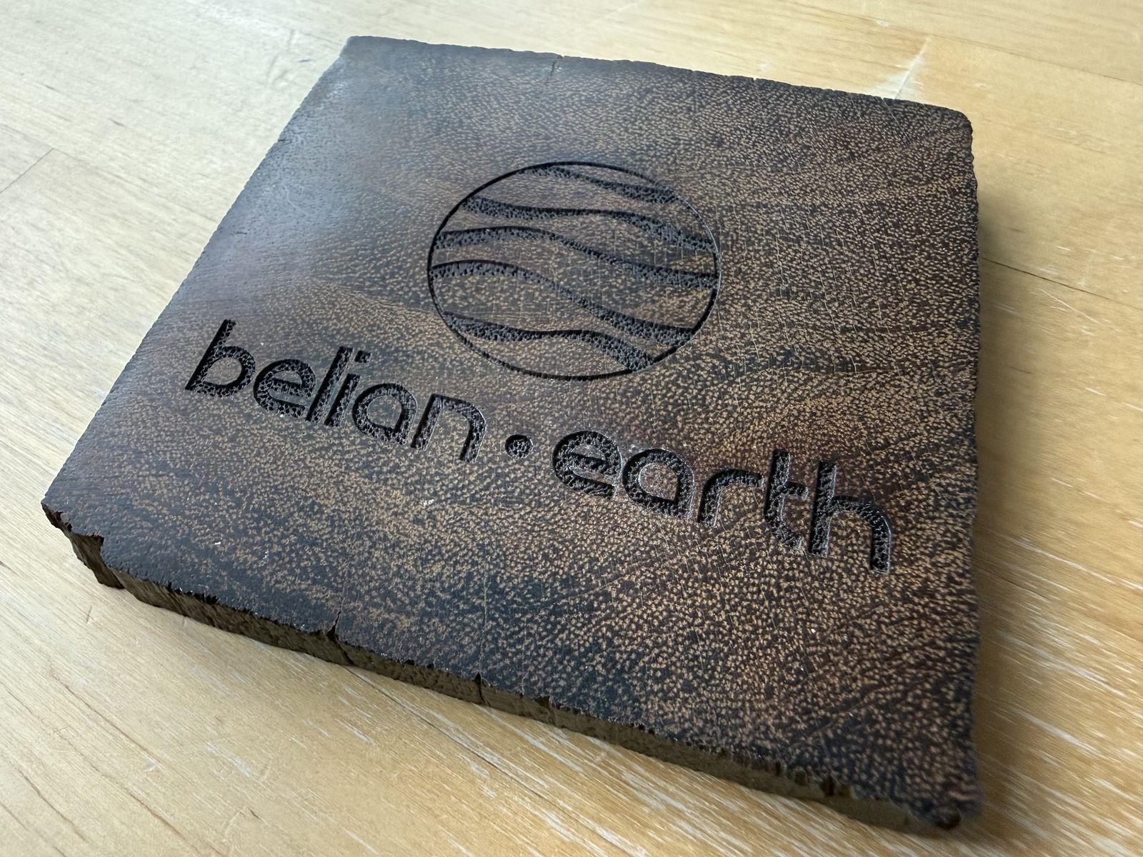 A square coaster of dark reclaimed belian wood on a pale wooden table, with belian.earth and the company mark burned into the surface and a rough sawn edge showing its thickness